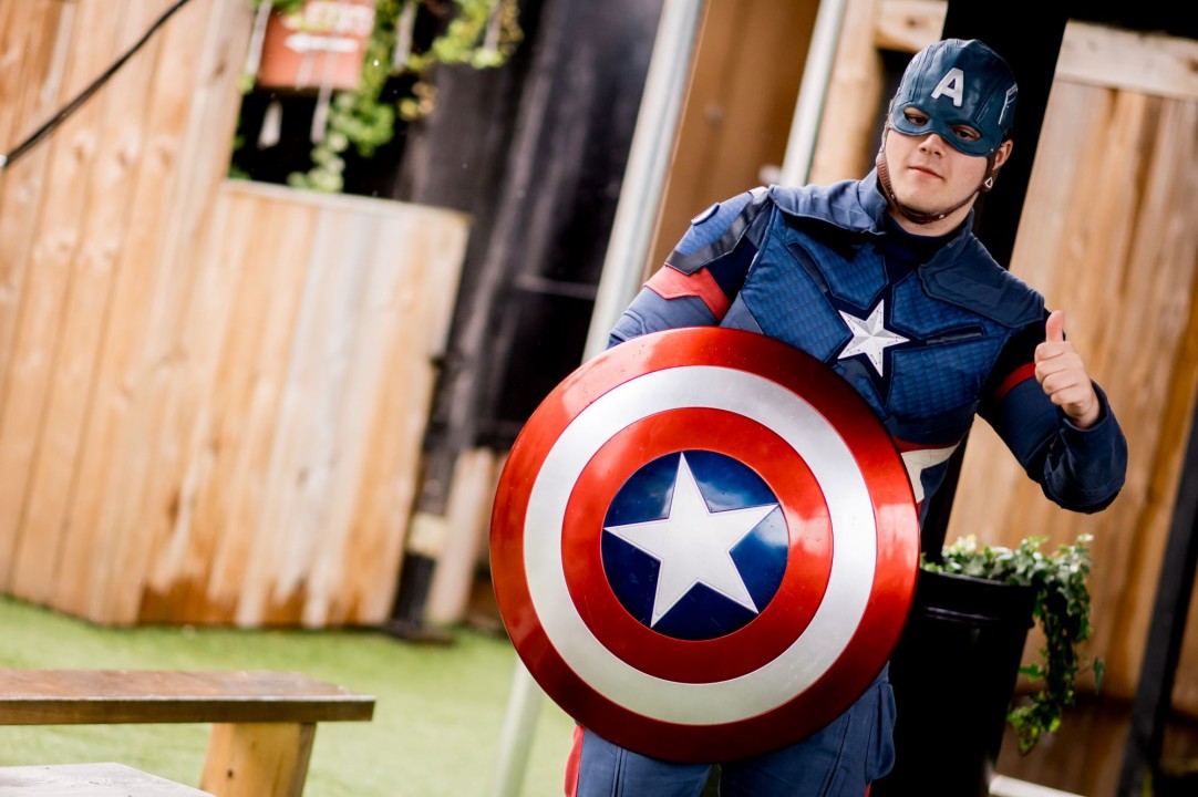 A man dressed in a blue superhero costume holding a round red, white and blue shield with a star in the middle