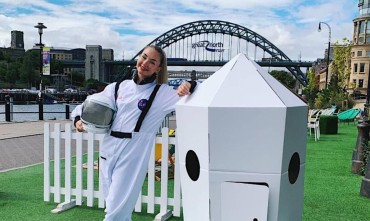 A woman dressed in an astronaut costume next to a cardboard model of a spaceship